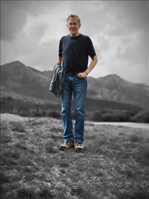 A man in blue jeans with a sweatshirt draped over his arm, and his hands in his pockets, stands in a grassy clearing with rolling hills in the background. 